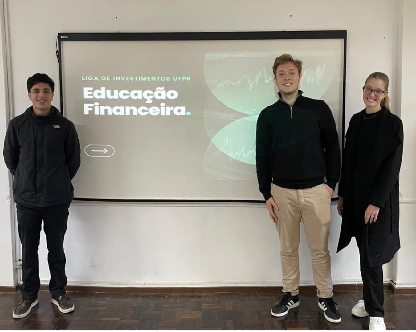 Três membros da Liga ao lado da tela com o slide Educação Financeira