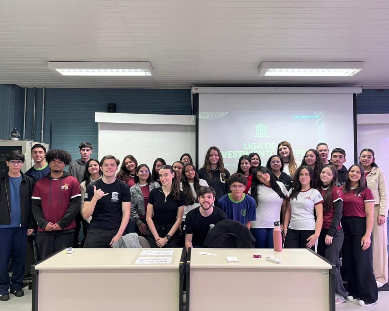 Turma de estudantes e membros da Liga em sala de aula, com a apresentação da Liga projetada ao fundo